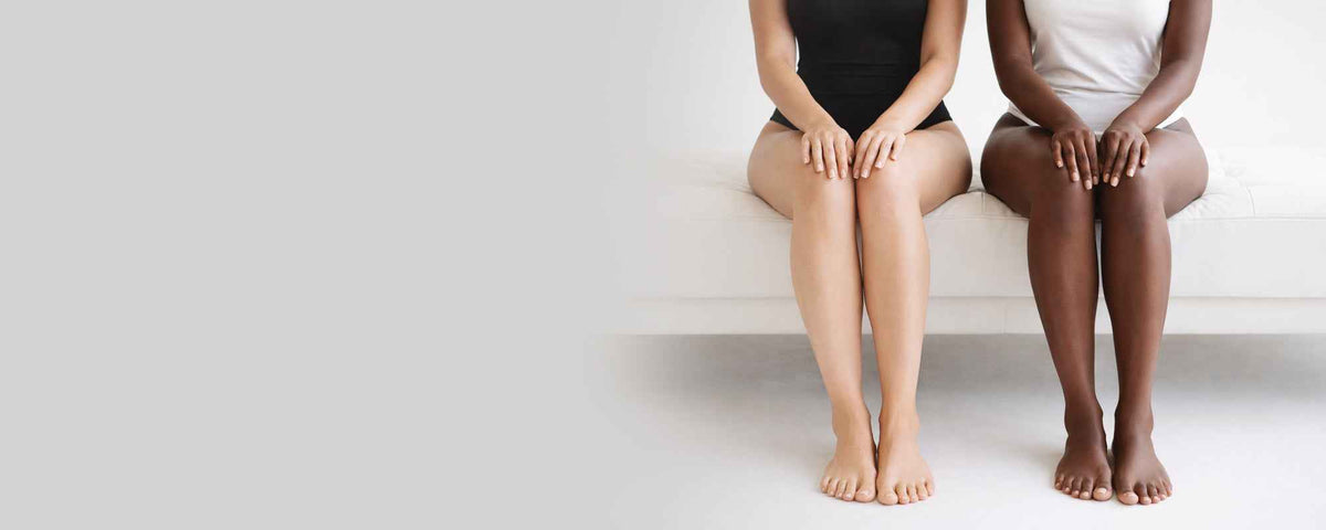 Two women with bare legs sitting on a white couch with a neutral background