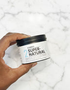 Hand holding a jar of Super-Natural body butter stretch mark product against a marble background